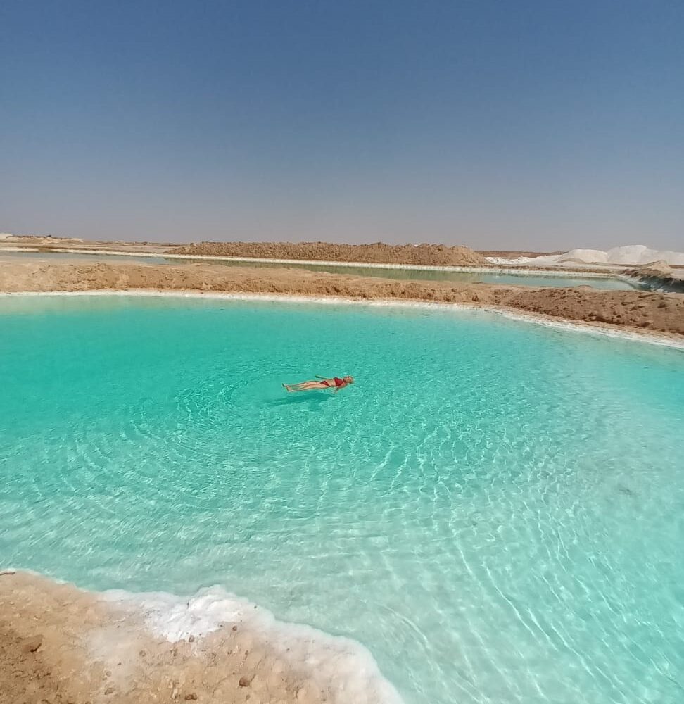 Siwa: La Magia de Flotar en el Agua de Sal Una persona flotando sin esfuerzo en la superficie de un lago salado de color turquesa en el Oasis de Siwa, con depósitos de sal cristalina en las orillas