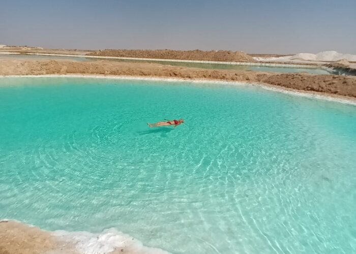 Una persona flotando sin esfuerzo en la superficie de un lago salado de color turquesa en el Oasis de Siwa, con depósitos de sal cristalina en las orillas