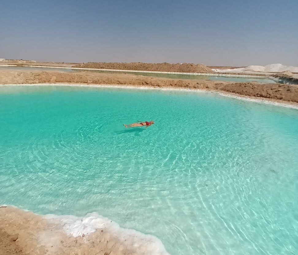 Una persona flotando sin esfuerzo en la superficie de un lago salado de color turquesa en el Oasis de Siwa, con depósitos de sal cristalina en las orillas