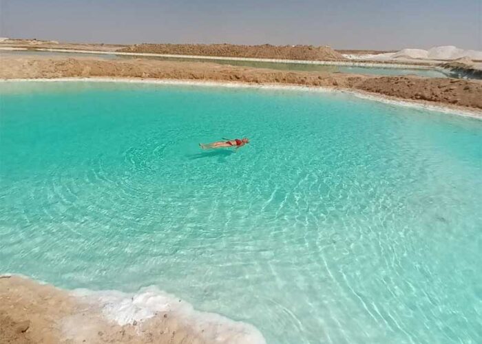 Une personne flottant sans effort dans les eaux turquoise et très salées d'un lac de l'Oasis de Siwa, avec des dépôts de sel blanc