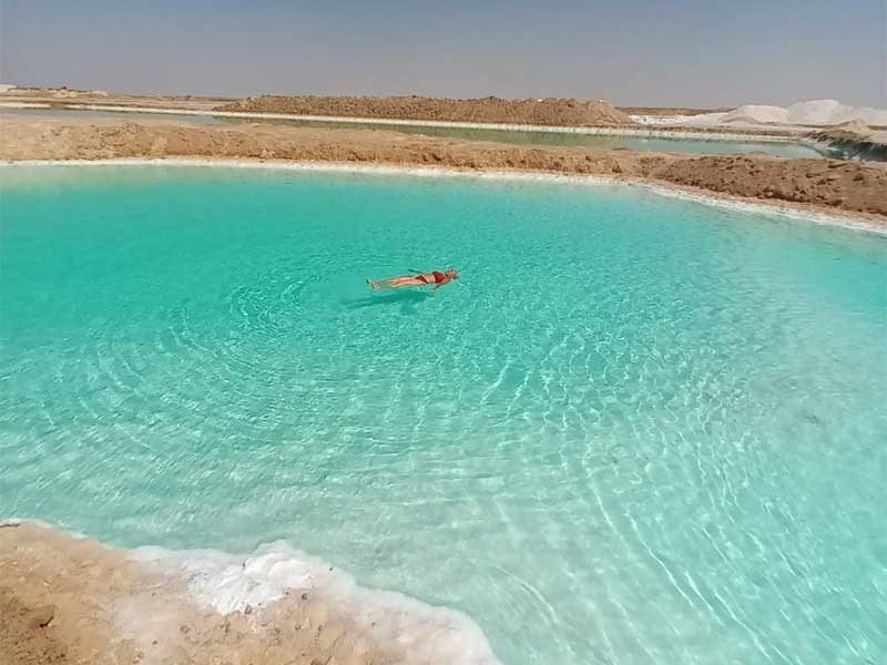 Une personne flottant sans effort dans les eaux turquoise et très salées d'un lac de l'Oasis de Siwa, avec des dépôts de sel blanc