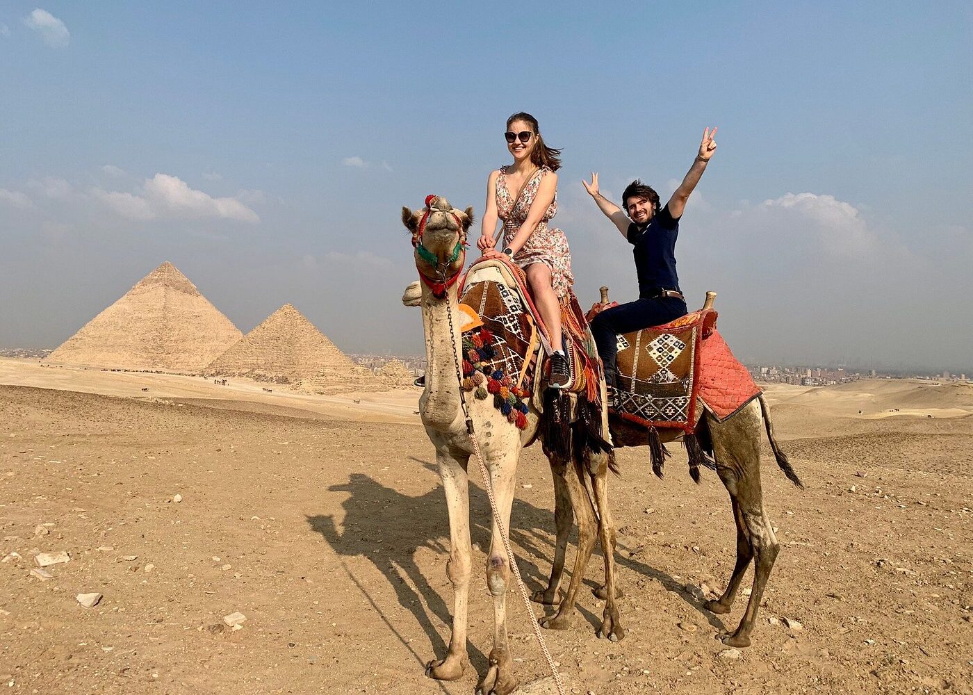 Gizé: Aventura em Camelo Um casal sorridente a cavalgar camelos pelo deserto de Gizé, com as majestosas Pirâmides ao fundo sob um céu azul