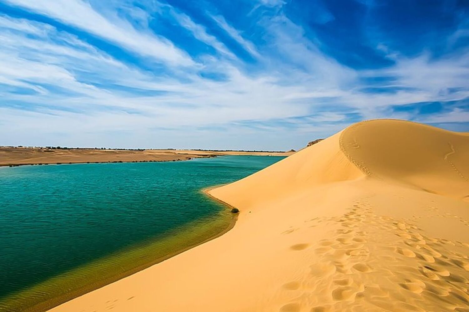 Superfície calma do Lago Mágico em Fayoum, refletindo cores variadas do céu e do deserto, com dunas ao redor